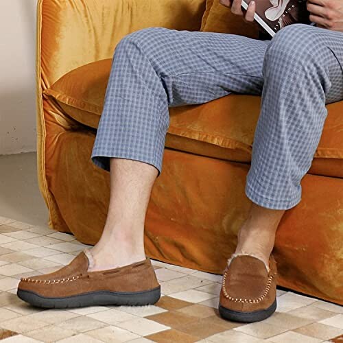 Person sitting on a couch wearing brown loafers and plaid pants, holding a book, enjoying comfort at home